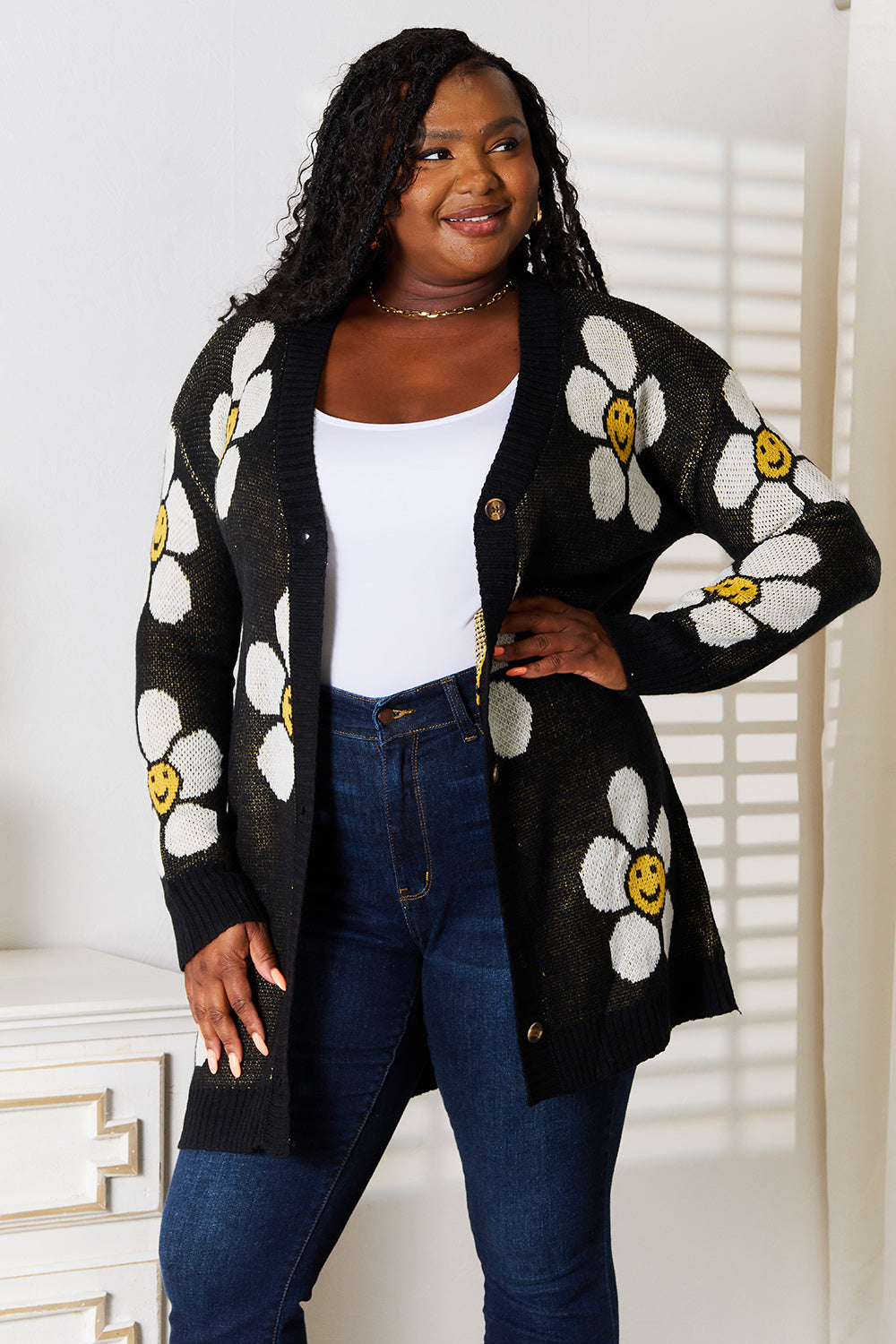 A woman stands indoors wearing the Perfee Floral Button Down Longline Cardigan, which features stylish black fabric adorned with white and yellow smiley face flowers. She pairs it with a white top and blue jeans, looking off to the side while smiling.