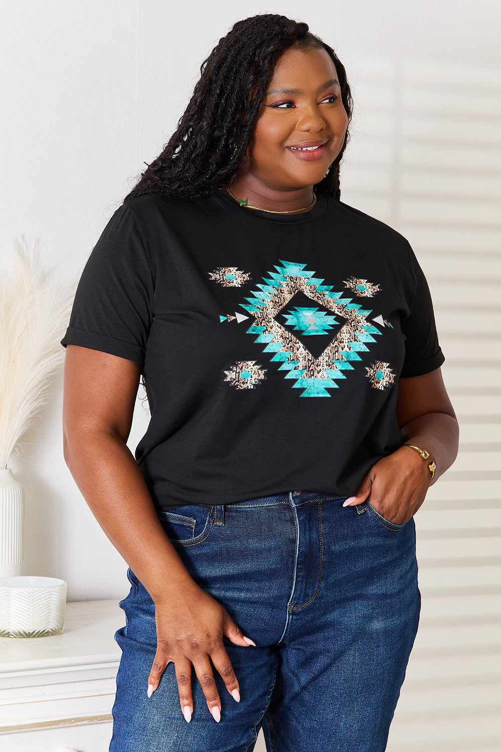 A woman stands indoors wearing the Simply Love Graphic Short Sleeve T-Shirt, a stylish black tee featuring a striking blue and white geometric pattern. Made from comfortable fabric, she has one hand on her hip and is smiling.