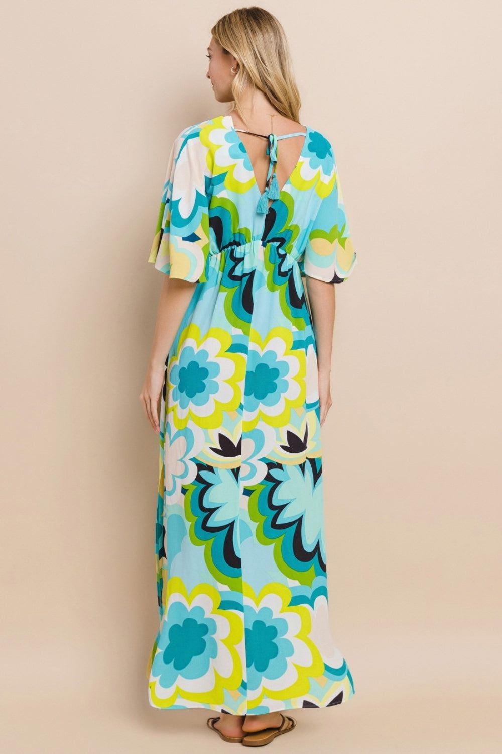 Against a simple backdrop, a woman models the ODDI Floral Printed Slit Maxi Dress, exuding a sense of versatile elegance. This maxi dress embodies summer chic with its vibrant floral design featuring shades of blue, green, and yellow.