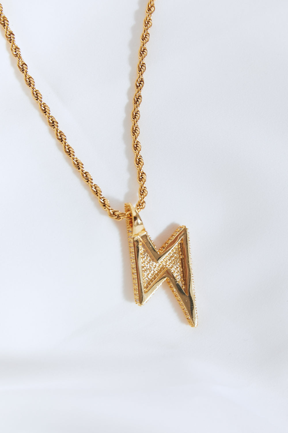 A Brass Lightning Pendant Necklace, featuring a gold twisted chain and a lightning bolt-shaped pendant encrusted with small cubic zirconia stones, displayed on a light blue background.