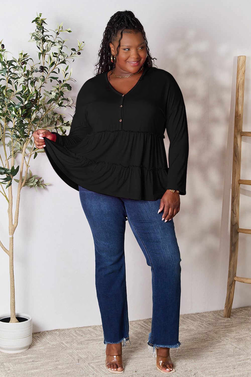 Person stands indoors near a plant, wearing the Double Take Half Button Long Sleeve Ruffle Hem Blouse in gray and blue jeans, smiling while looking to the side. The slightly stretchy fabric of the blouse adds comfort while its buttoned design offers a touch of elegance.