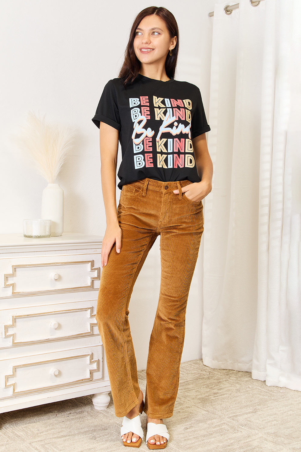 A woman stands with her hands in the pockets of her brown corduroy pants, wearing a casual Simply Love BE KIND Graphic Round Neck T-Shirt that reads "BE KIND" in large colorful letters, showcasing positive message clothing.