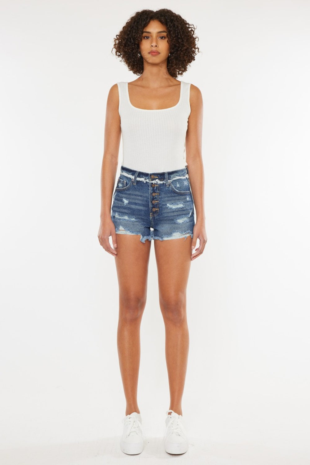 A woman with curly hair wearing a white sleeveless top and Kancan Distressed Button Fly Denim Shorts stands against a plain background, perfect for a stylish summer wardrobe.