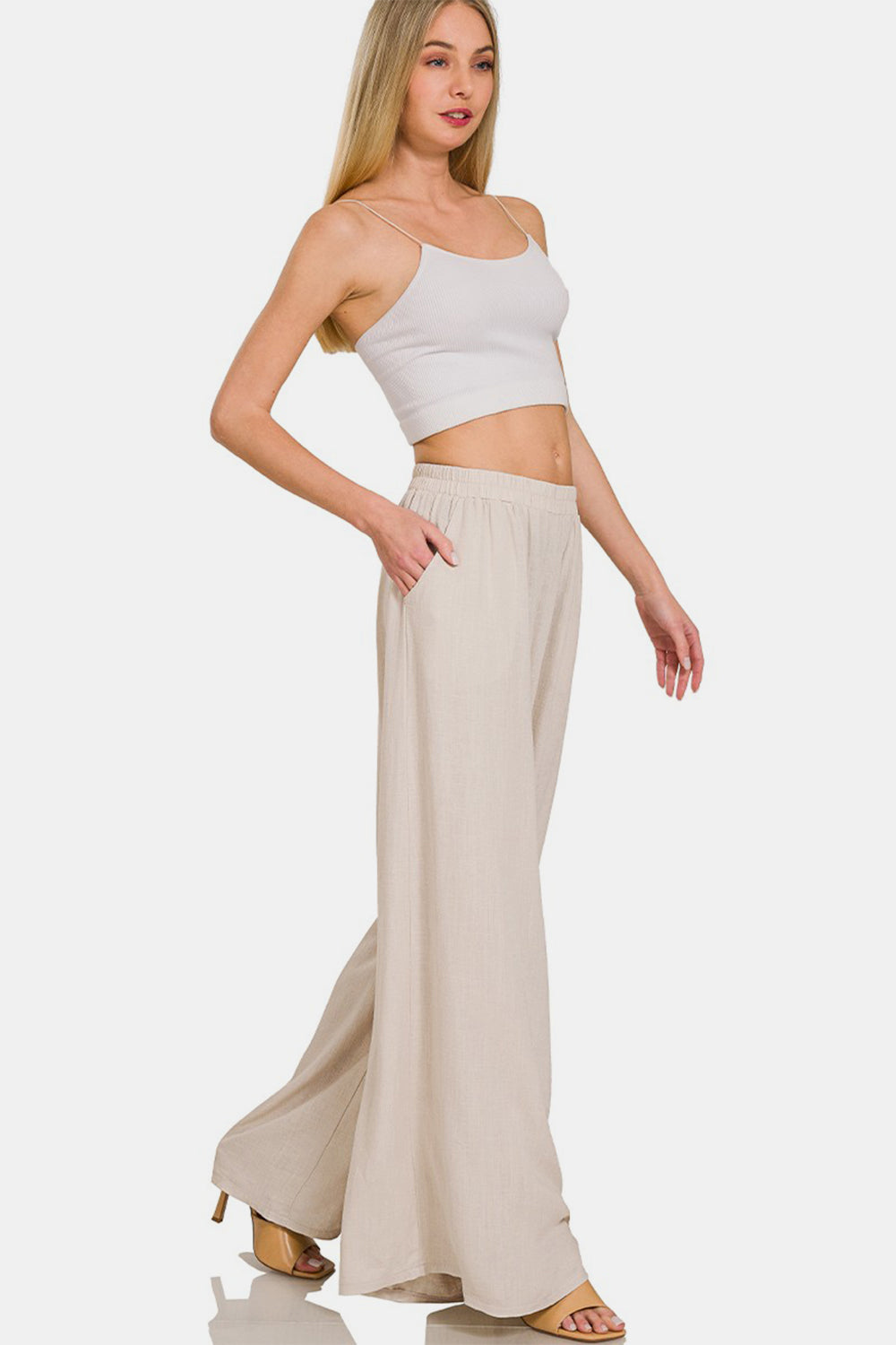 Person wearing Zenana Pleated Linen Blend Wide Leg Pants in beige with hands in pockets, shown from waist to feet. The pleated pants have an elastic waistband and flowy fabric. Model is also wearing light-colored open-toed sandals.