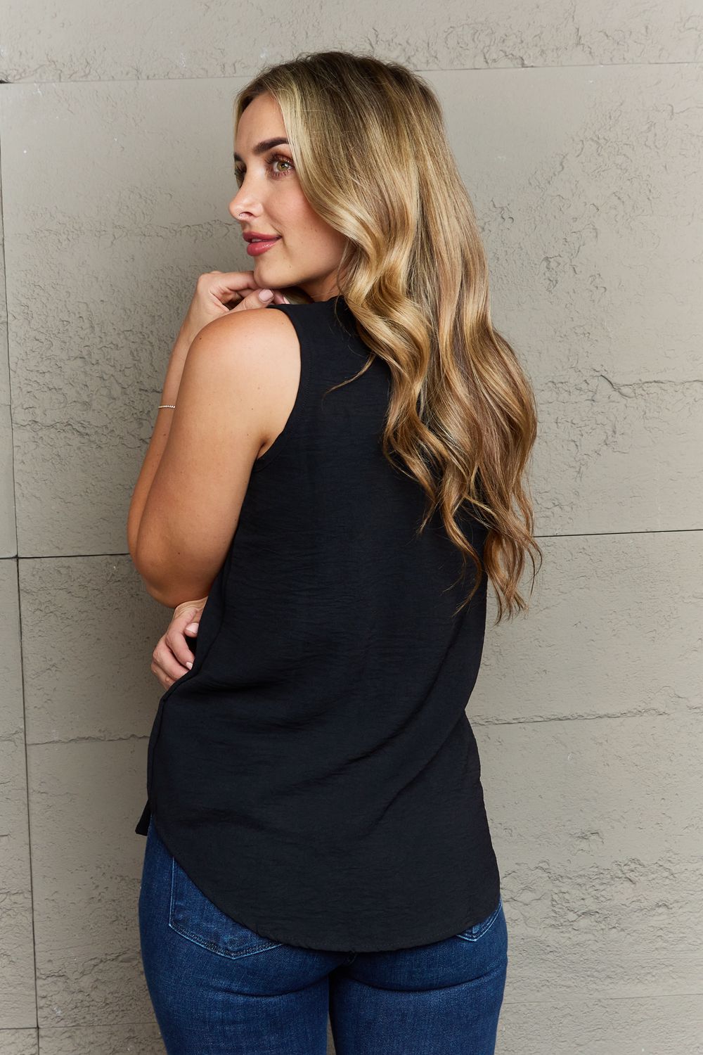 A woman with long blonde hair stands against a textured wall, wearing the Ninexis First Glance Sleeveless Neckline Slit Top in black and blue jeans. Her left hand is on her hip while her right hand rests on the wall.