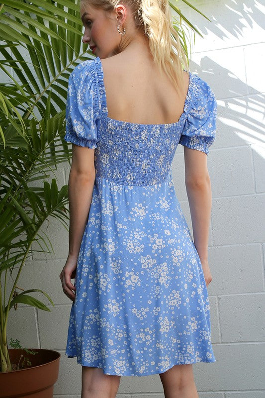 A person wearing the SS puff sleeve dress in a blue floral print stands in front of green palm leaves and a white wall.