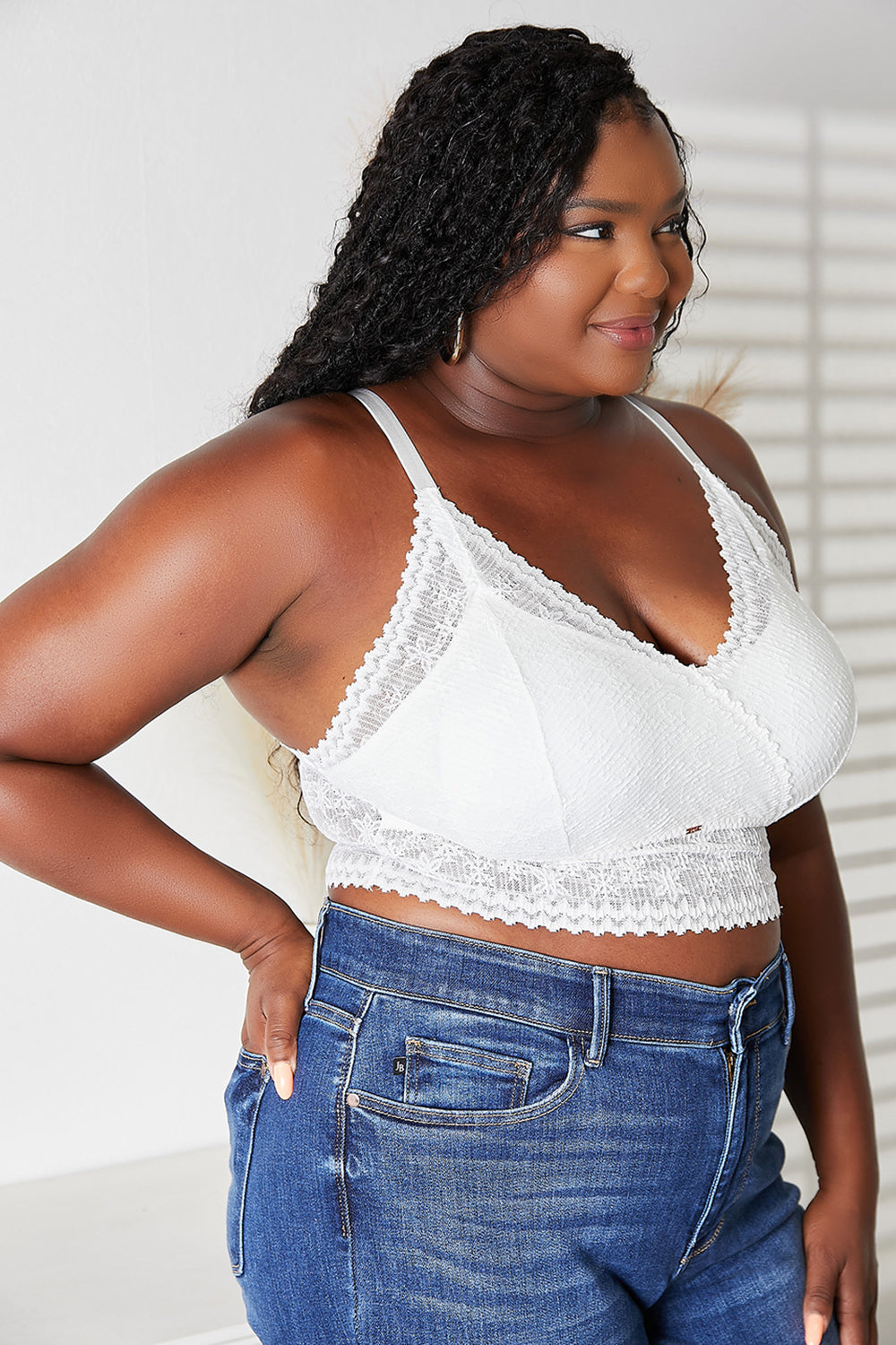 A woman wearing a JadyK Skye Full Size Lace Bralette with removable padding and blue jeans stands indoors, looking directly at the camera.