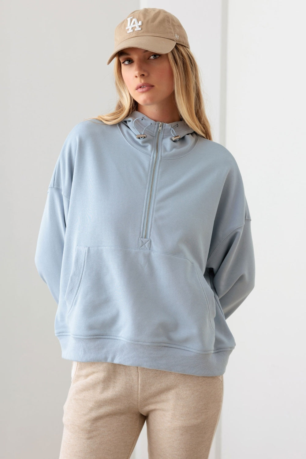 In a neutral-colored room, a person is wearing the Le Lis Half Zip Drawstring Mock Neck Hoodie in light blue along with a beige cap.