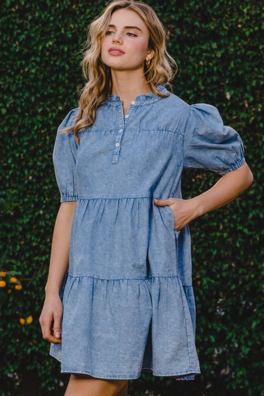 A person wearing the ODDI Washed Tiered Mini Denim Dress with puffed sleeves stands in front of a green leafy background.