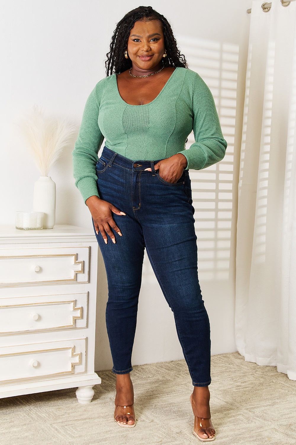 A person wearing a fashion-forward Basic Bae Scoop Neck Long Sleeve Top in green and dark jeans stands indoors, smiling and looking to the side.