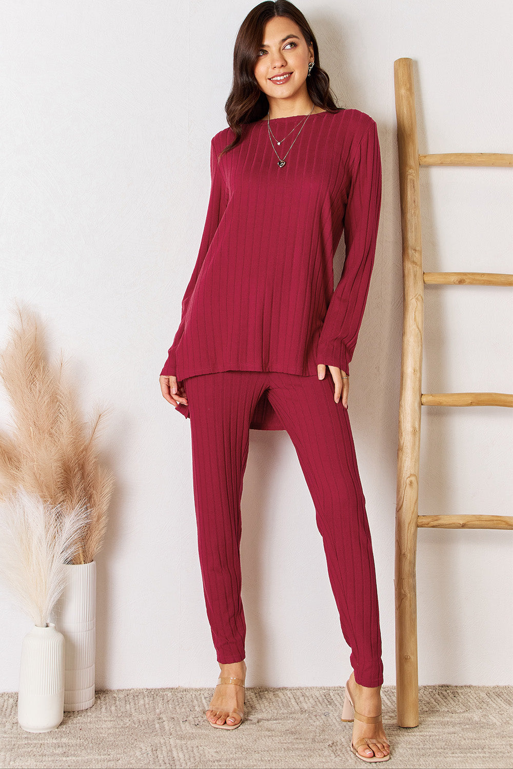 A woman in a Basic Bae Full Size Ribbed Round Neck High-Low Slit Top and Pants Set poses indoors near a wooden ladder. She is wearing the matching maroon slightly stretchy top and pants, paired with beige heels. Pampas grass in a white vase is visible in the foreground. For care, machine wash cold.