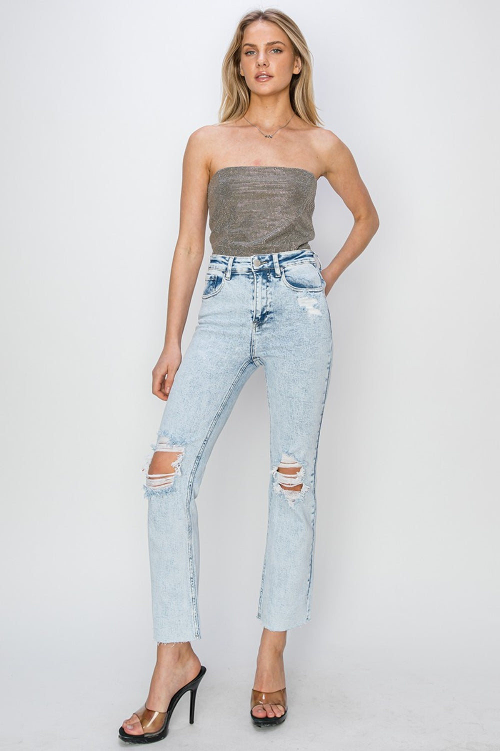 Person wearing RISEN High Rise Distressed Ankle Jeans in light blue with rips and raw hems, paired with transparent-strap high heels.