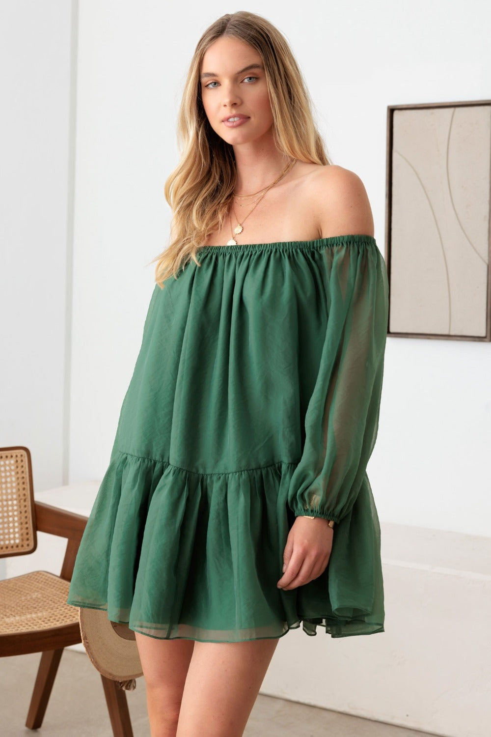 A woman stands indoors near a chair and a framed artwork on the wall, wearing the stunning Le Lis Organza Off Shoulder Puff Sleeve Mini Dress in green. This off-the-shoulder dress with long sleeves makes for a perfect special occasion ensemble.