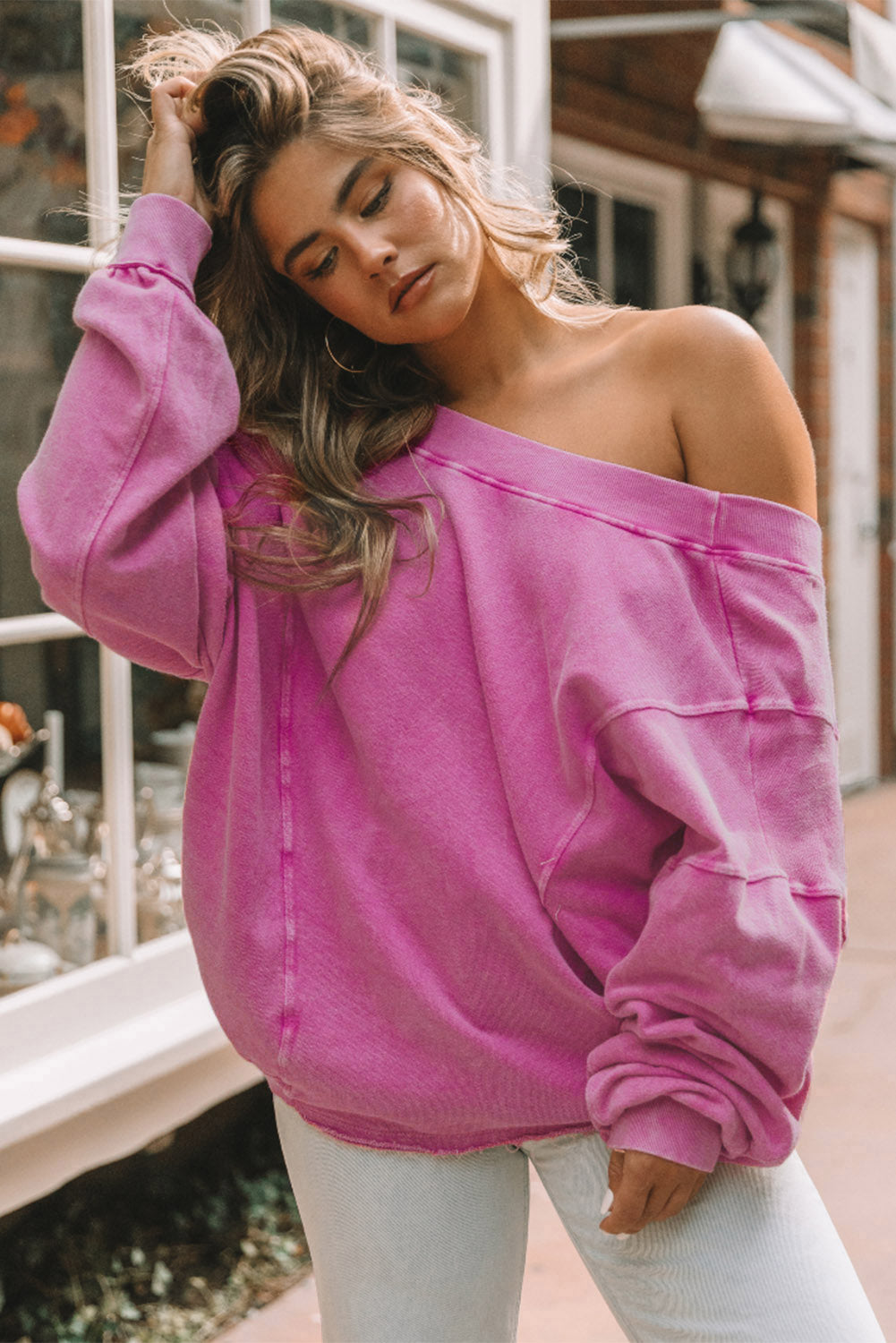 A woman stands with her back to the camera, wearing a long-sleeved Rose Exposed Seam Twist Open Back Oversized Sweatshirt and light blue jeans, showcasing a relaxed fit.