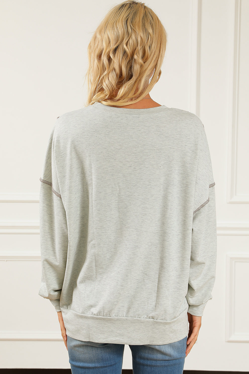 A person with wavy blonde hair is shown from the back, standing in front of a white paneled wall, wearing a light gray Asymmetrical Leopard Texture Splicing Loose Sweatshirt and blue jeans.