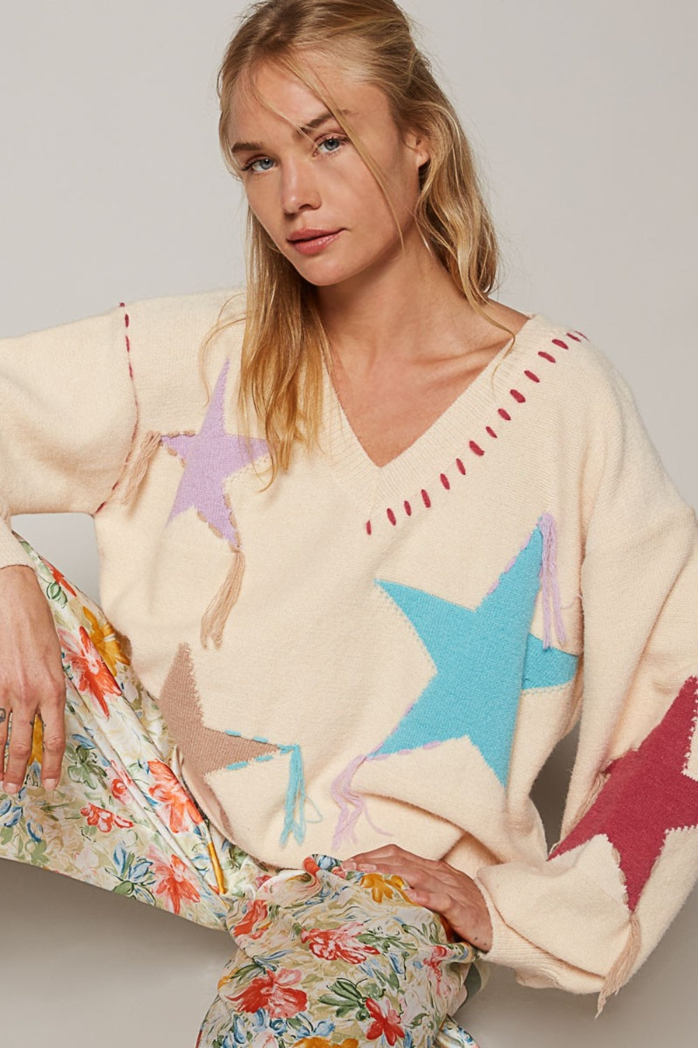 Person wearing a cozy POL V-Neck Long Sleeve Star Fringe Sweater and floral pants, sitting against a plain background.