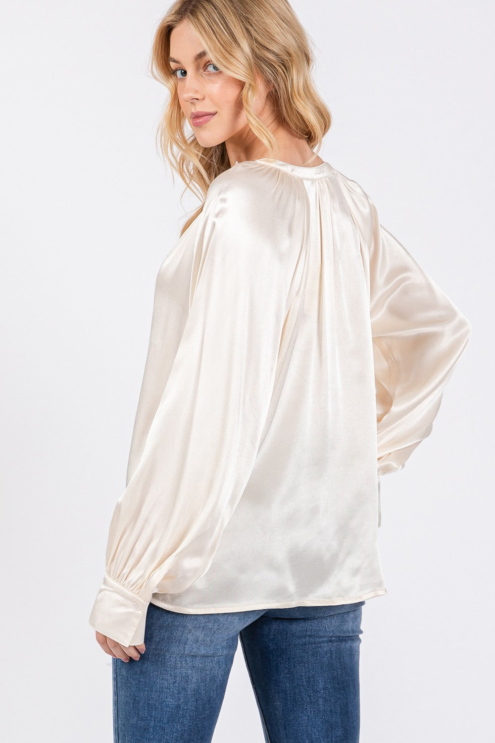 A person is wearing a SAGE + FIG Notched Long Sleeve Blouse in cream-colored satin paired with blue jeans.