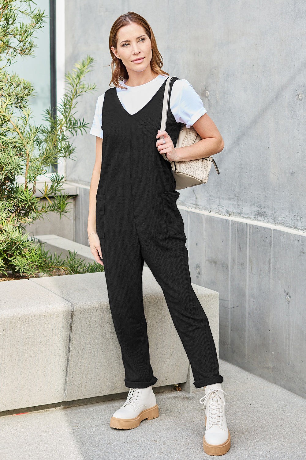 A person stands outdoors wearing the Double Take Full Size Sleeveless Straight Jumpsuit in blue and brown sandals, holding a cream-colored bag. In the background, there's a wooden bench and palm trees.