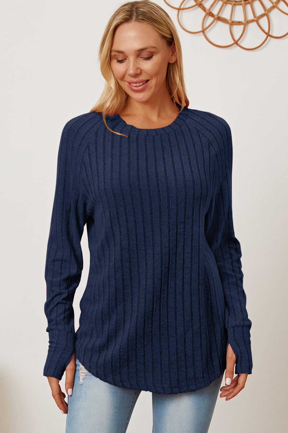 A person wearing the Basic Bae Full Size Ribbed Thumbhole Sleeve T-Shirt in blue and light-washed jeans stands smiling, with a decorative wall hanging in the background.