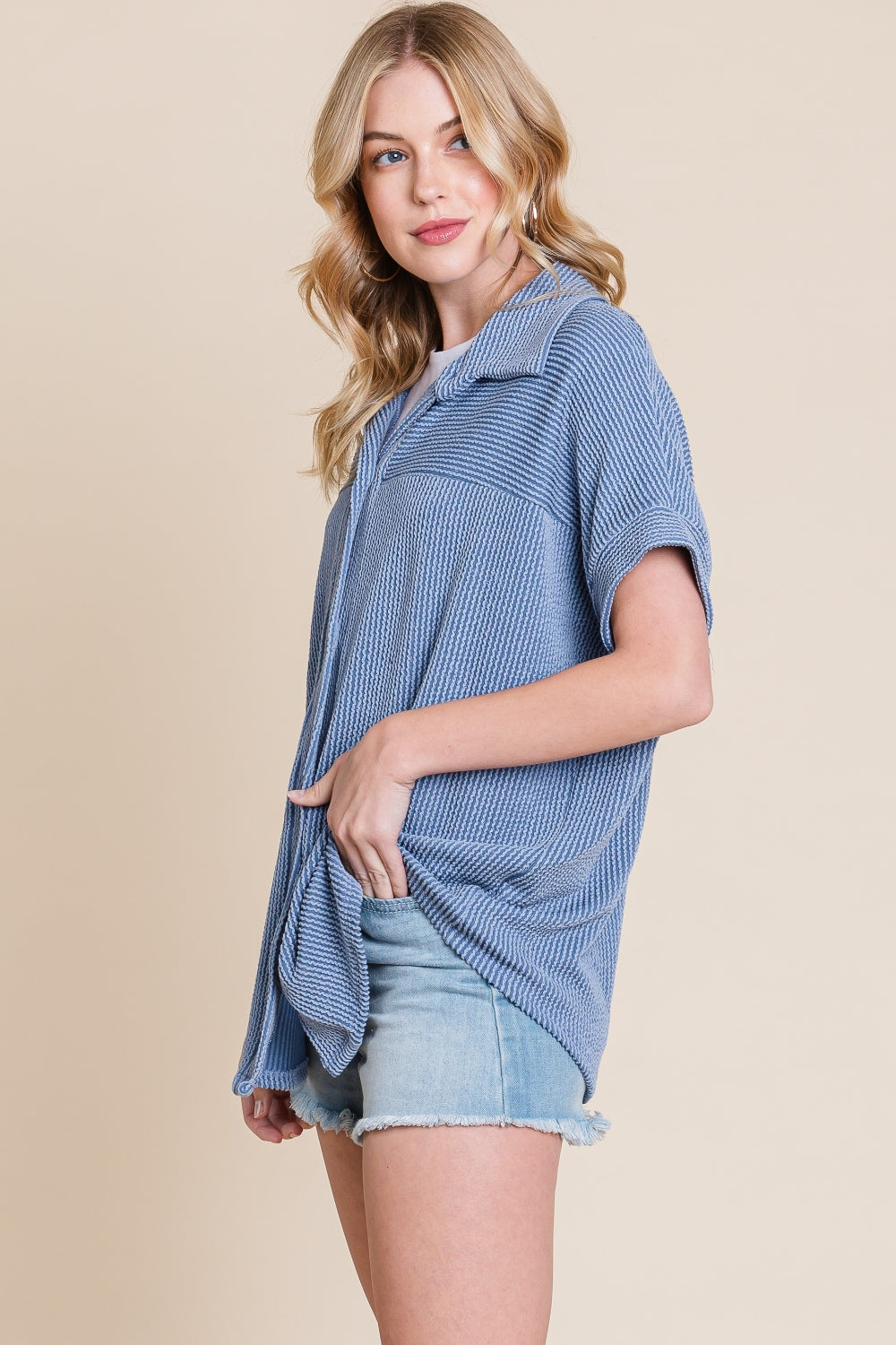 A woman wearing a BOMBOM Button Up Short Sleeves Ribbed Shirt in blue over a white shirt, paired with light denim shorts, stands against a beige background.