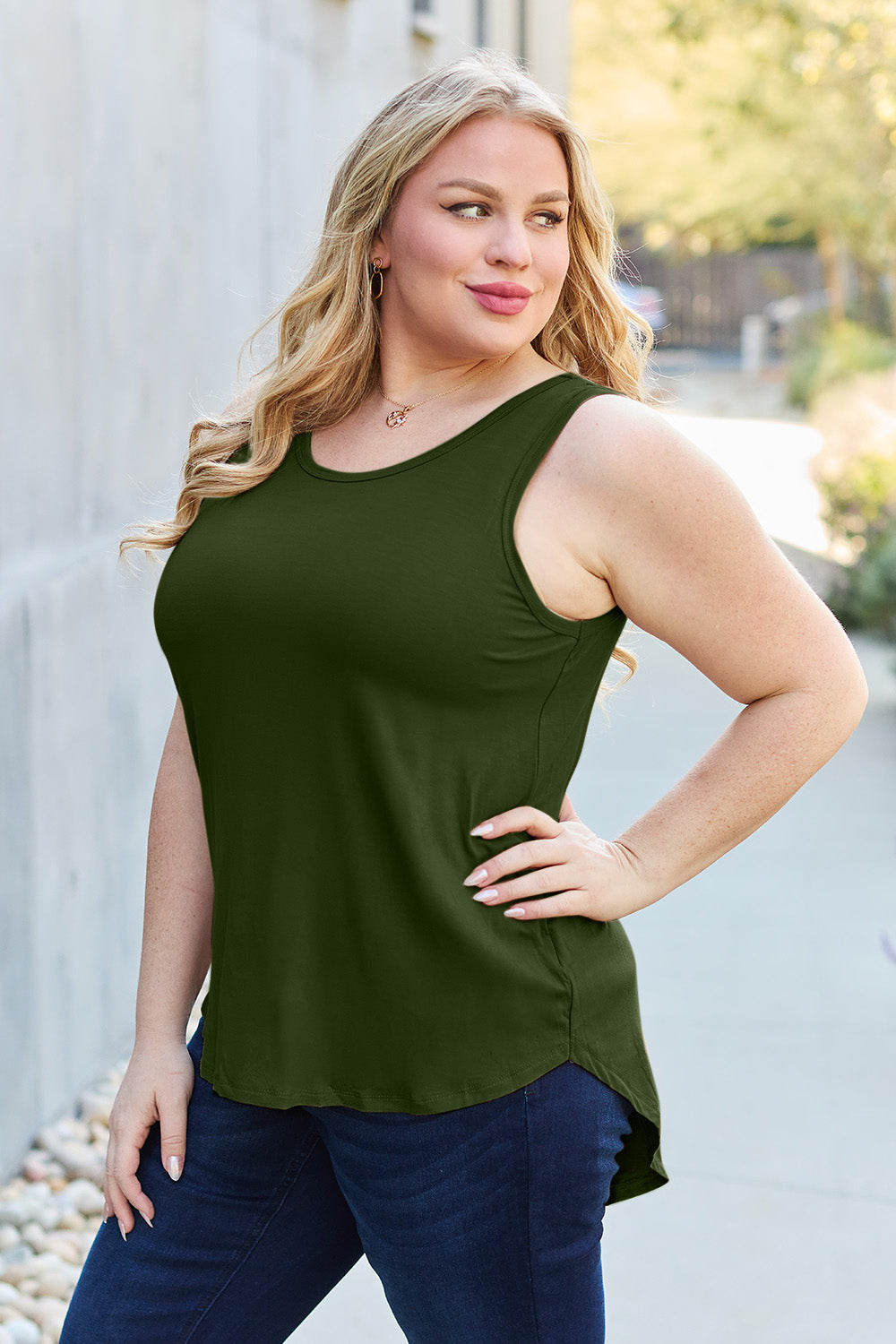 A woman wearing sunglasses, blue jeans, and the Basic Bae Full Size Round Neck Curved Hem Tank is standing in front of a concrete wall, holding her glasses with one hand and carrying a beige shoulder bag. Her basic style complements the slightly stretchy fabric of her outfit, ensuring comfort while maintaining chic simplicity.