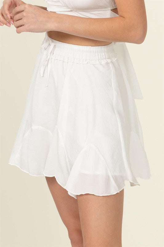 A person is partially visible in the image wearing the Not Your Girl Drawstring Ruffled Mini Skirt, featuring a bright pink flared silhouette with an elastic waistband and drawstring, paired with a matching top.