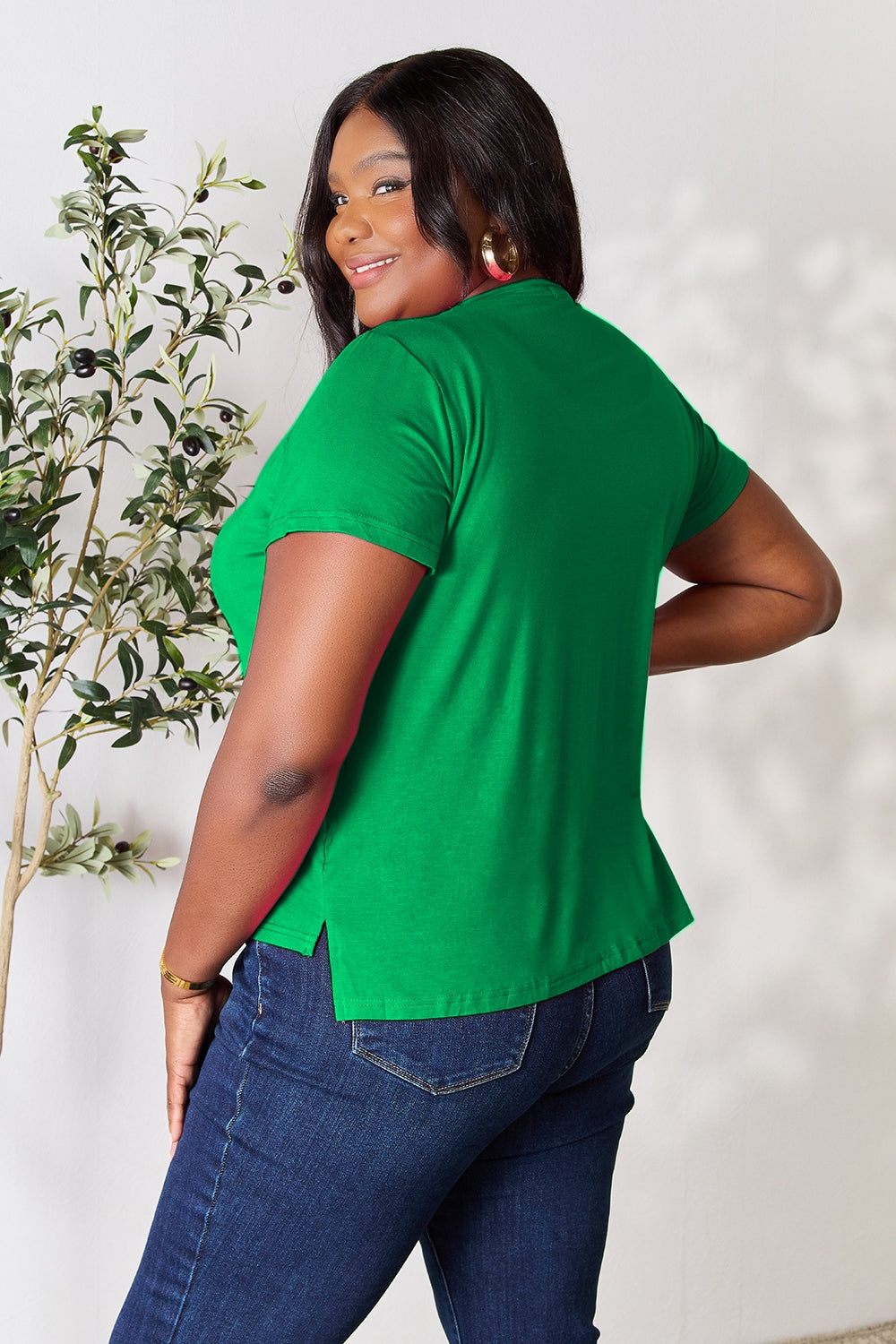 A person with wavy hair, wearing the Basic Bae Full Size Round Neck Short Sleeve T-Shirt in bright pink and beige pants, stands near a plant, smiling and looking to the side. The outfit embodies a basic style and offers slightly stretchy comfort, ideal for those who value effortless elegance.