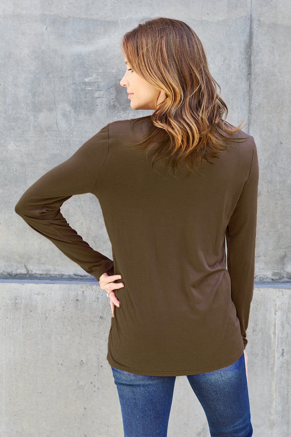 A woman with shoulder-length hair wearing the Basic Bae Full Size Round Neck Long Sleeve Top and blue jeans stands in front of a concrete wall, holding a beige handbag over her shoulder. Her outfit exemplifies basic style, with the top made from 100% polyester and recommended for machine wash cold.