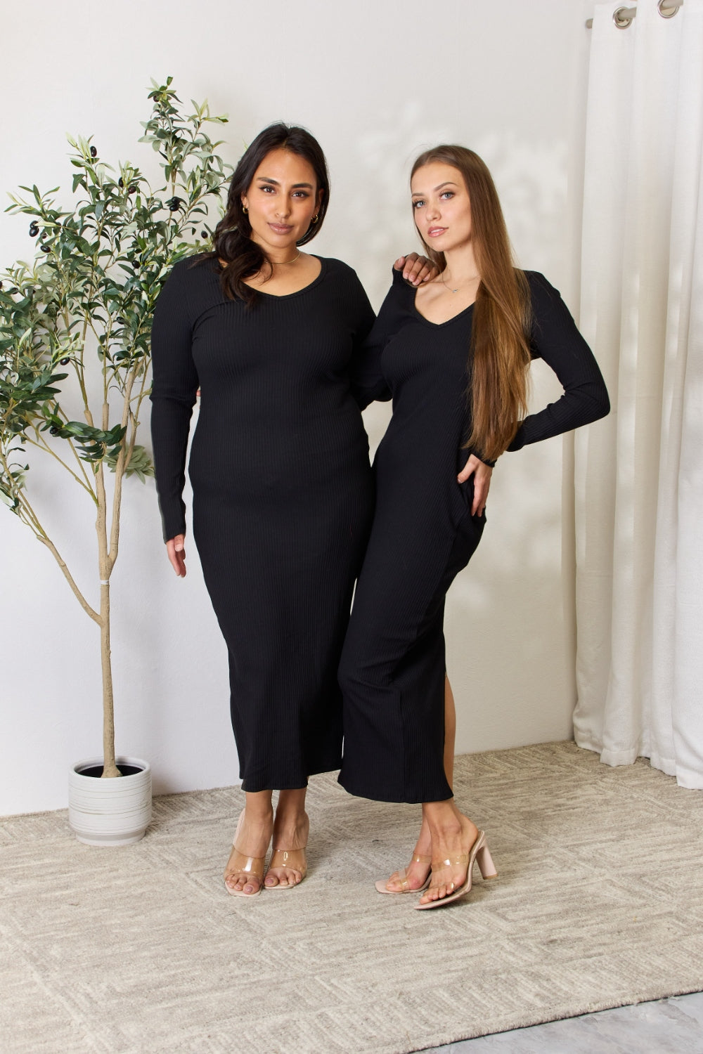 A person stands indoors wearing the chic Culture Code Full Size Ribbed Long Sleeve Midi Slit Dress paired with sandals, next to a potted plant.