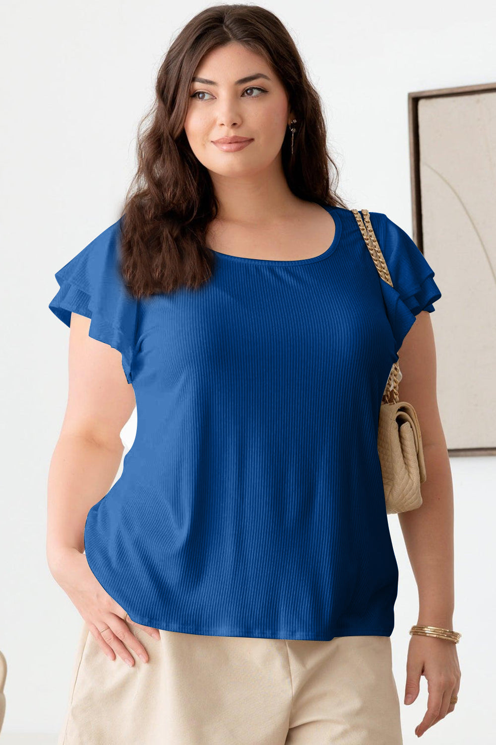 A person wearing the Gilli Plus Size Short Fluttery Sleeve Round Neck Top in blue and jeans stands indoors, showcasing an effortlessly chic style.