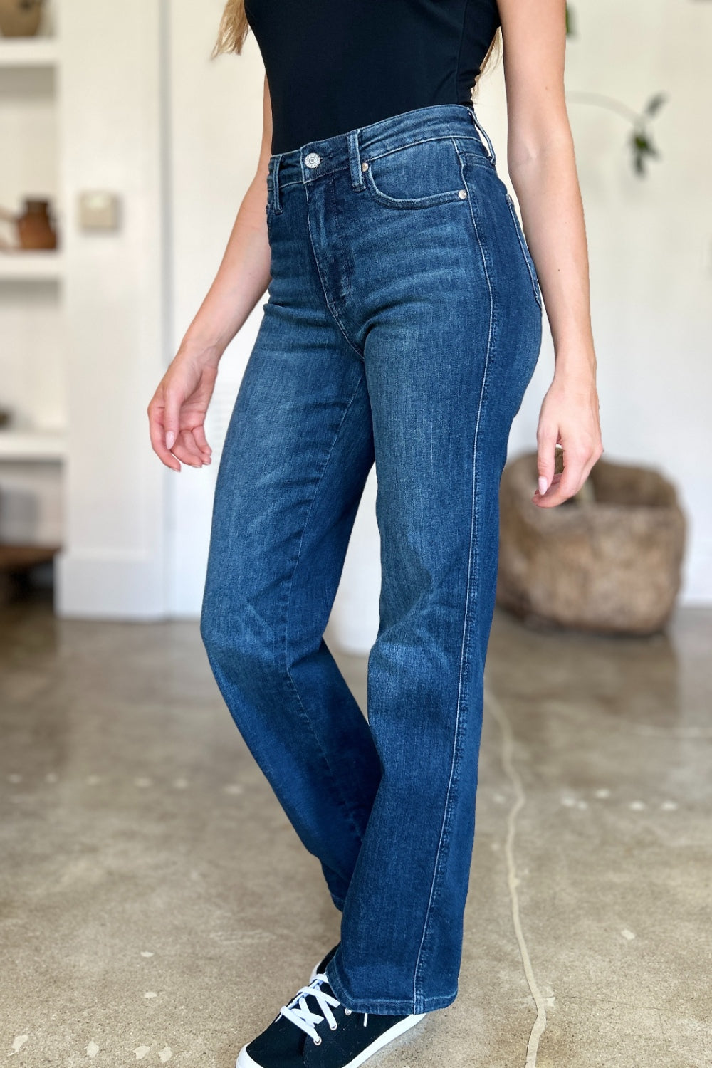 A person wearing a black top and Judy Blue Full Size Tummy Control Straight Jeans stands indoors with one hand on their hip. Their shoes are black with white soles. A plant and shelves are in the background.