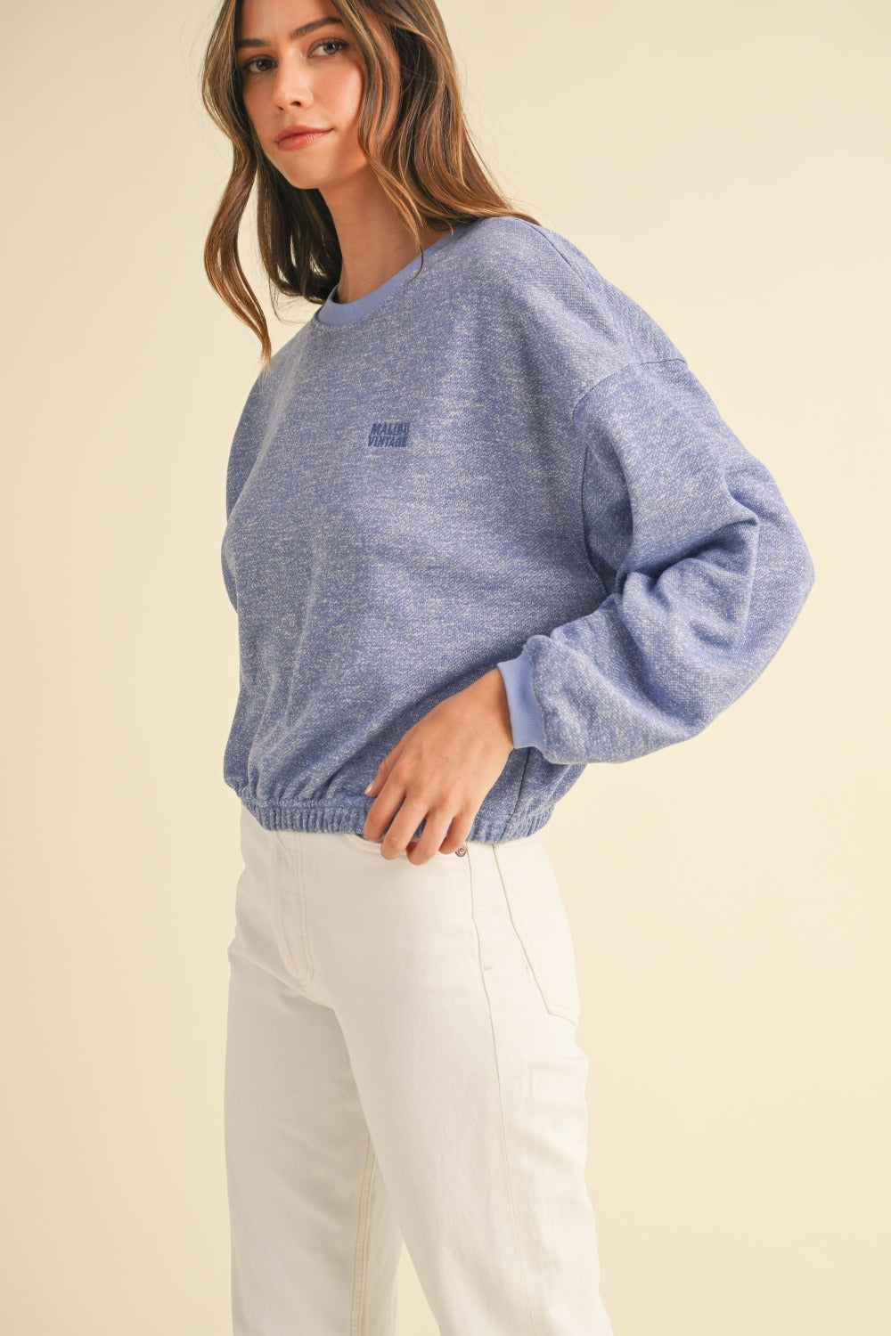 A person wearing a Mable Round Neck Letter Embroidery Crop Sweatshirt in blue, paired with white pants, stands against a plain background, touching the collar of the sweatshirt with one hand.