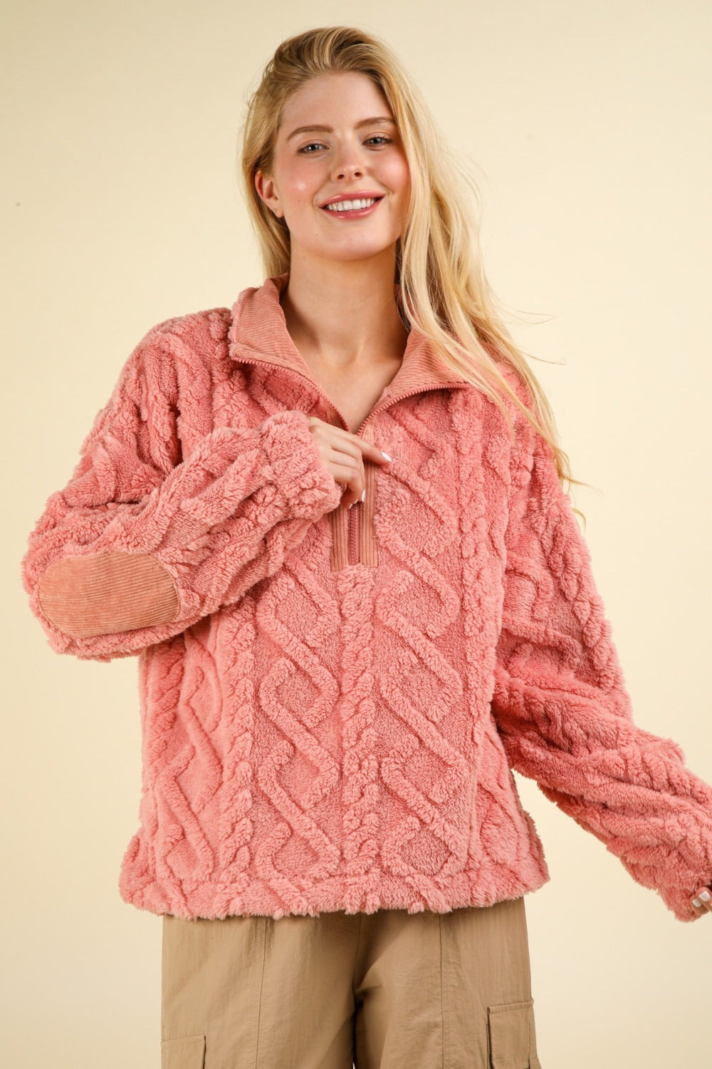 A person with long blonde hair wears a VERY J Fuzzy Fleece Half Zip Cable Pattern Sweatshirt in pink, paired with beige pants, standing against a plain light background. The sweatshirt is adorned with textured details and elbow corduroy patches.