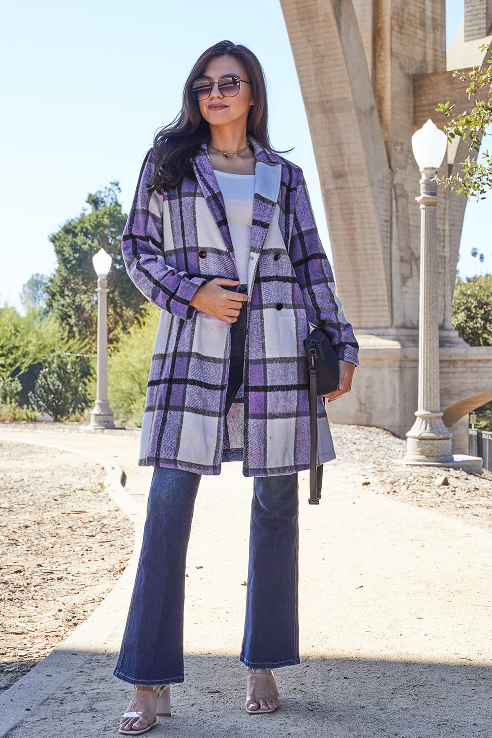 A woman stands on a sidewalk wearing the Double Take Full Size Plaid Button Up Lapel Collar Coat in pink and white over a white top and blue jeans, with her right hand lightly touching her hair. The polyester fabric of the coat makes it durable and perfect for an easy machine wash cold.