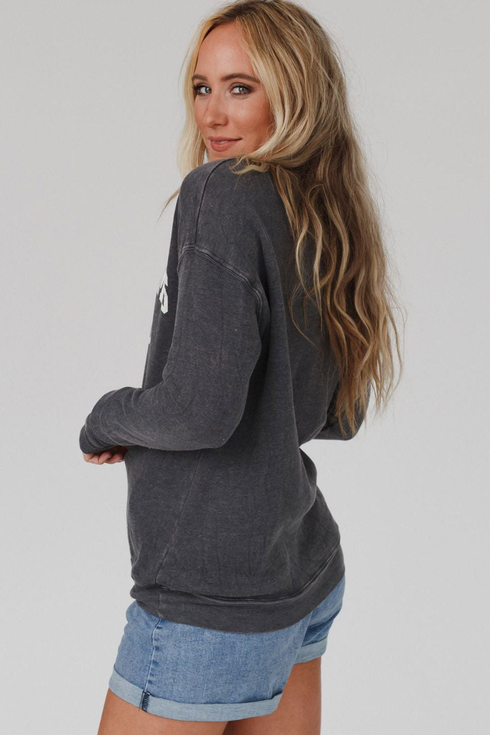 A person with long blond hair wearing a gray COOL MOMS CLUB drop shoulder sweatshirt and relaxed-fit blue jeans is seen from the back, standing near a pumpkin and a window.