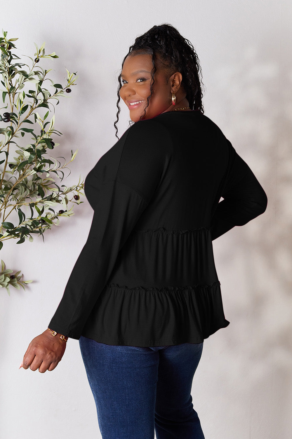 Person stands indoors near a plant, wearing the Double Take Half Button Long Sleeve Ruffle Hem Blouse in gray and blue jeans, smiling while looking to the side. The slightly stretchy fabric of the blouse adds comfort while its buttoned design offers a touch of elegance.