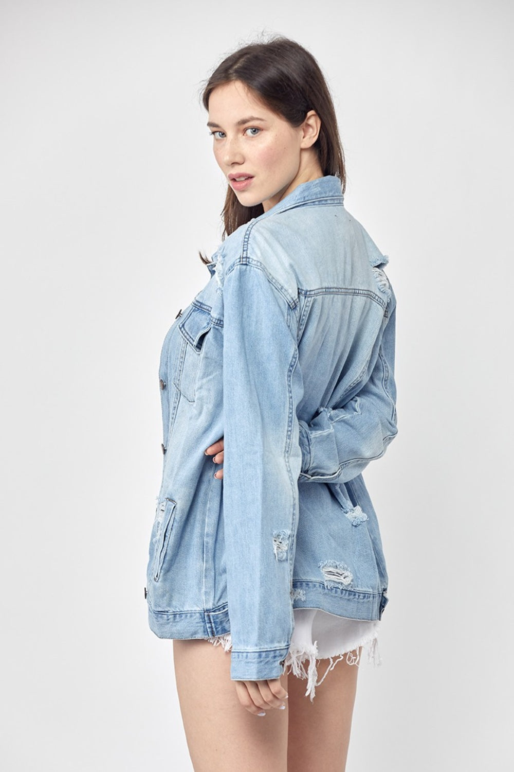 Dressed in the RISEN Full Size Distressed Long Sleeve Denim Jacket, a perfect wardrobe staple, this person effortlessly poses against a plain background while pairing it with a blue top and white shorts.