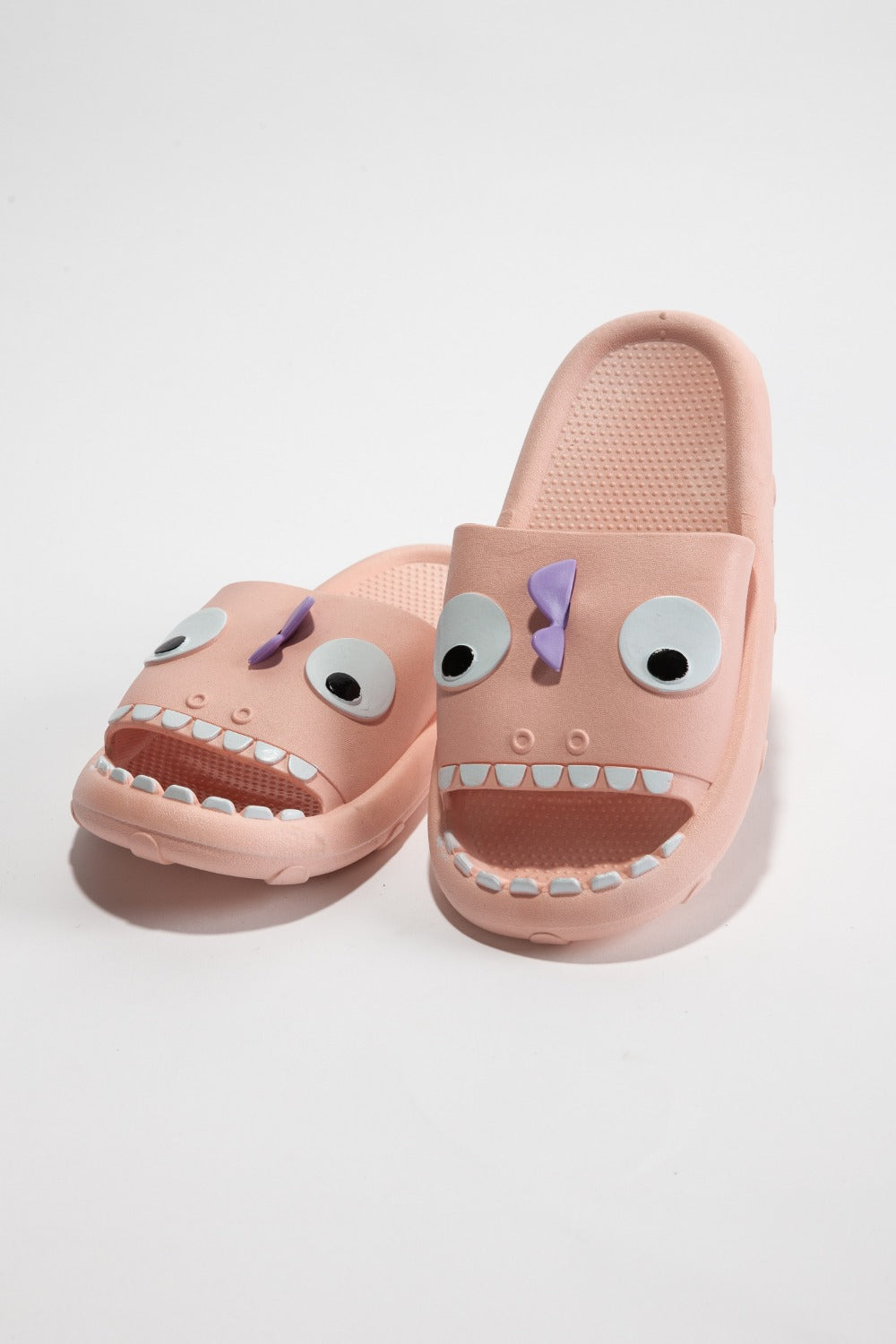 The NOOK JOI Monster Pillow Cloud Slides Non-Slip Slipper features cartoonish monster faces with large eyes, a purple nose, and an open mouth with teeth. Made of durable EVA material, these fun pink cloud slippers also boast an anti-slip design for added safety.