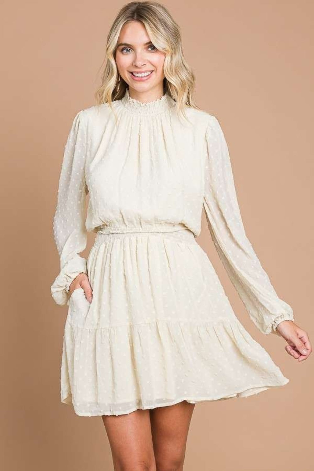 Wearing the Culture Code Swiss Dot Smock Neck Mini Dress with Pockets in light beige, a person stands against a brown backdrop, smiling, with one hand on the hip and the other gently holding the elegantly draped lightweight chiffon fabric.