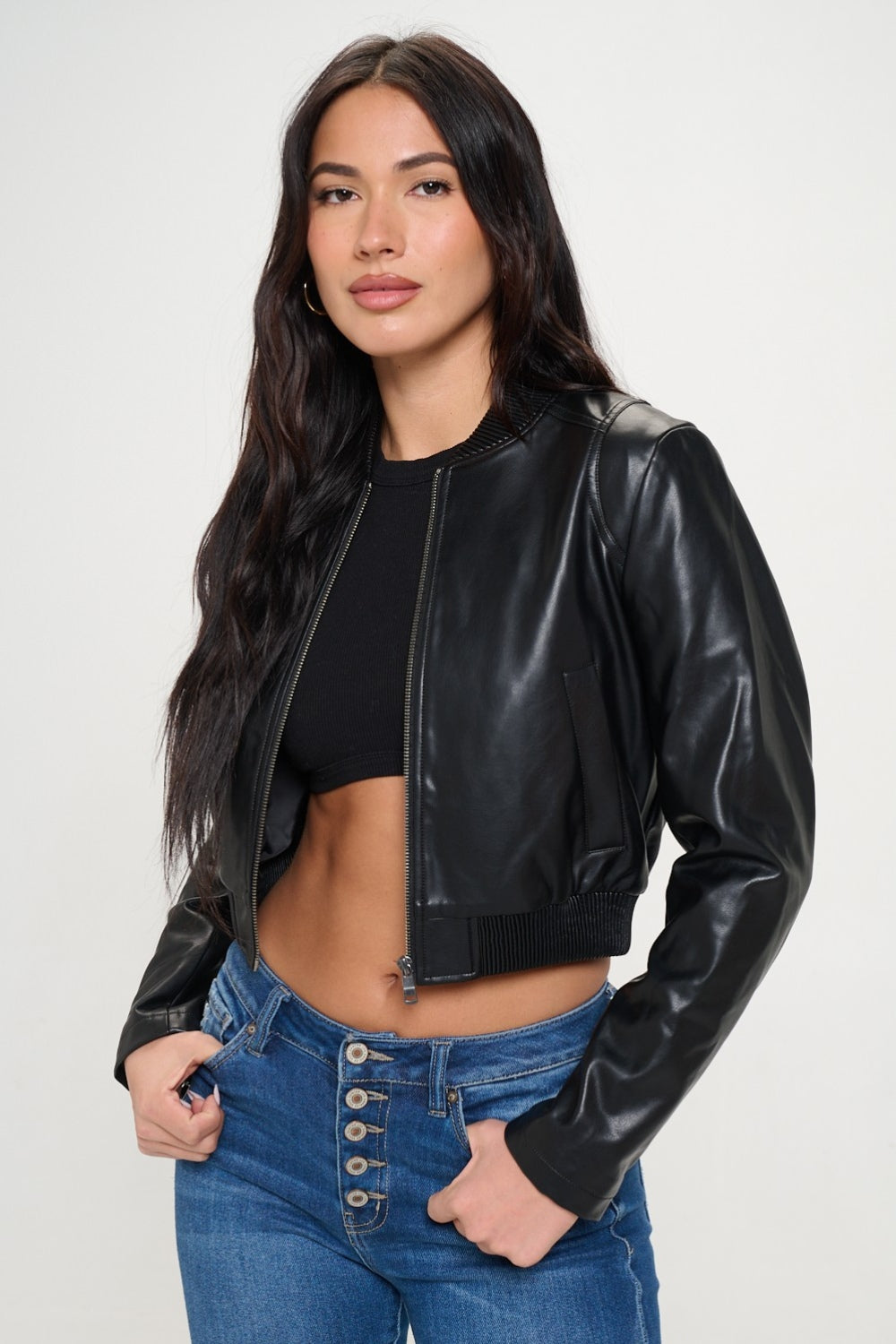 A woman wearing a Coalition LA Zip Up Cropped Bomber Jacket over a black crop top and blue jeans with buttons, looking at the camera with her hands in her jacket pockets.