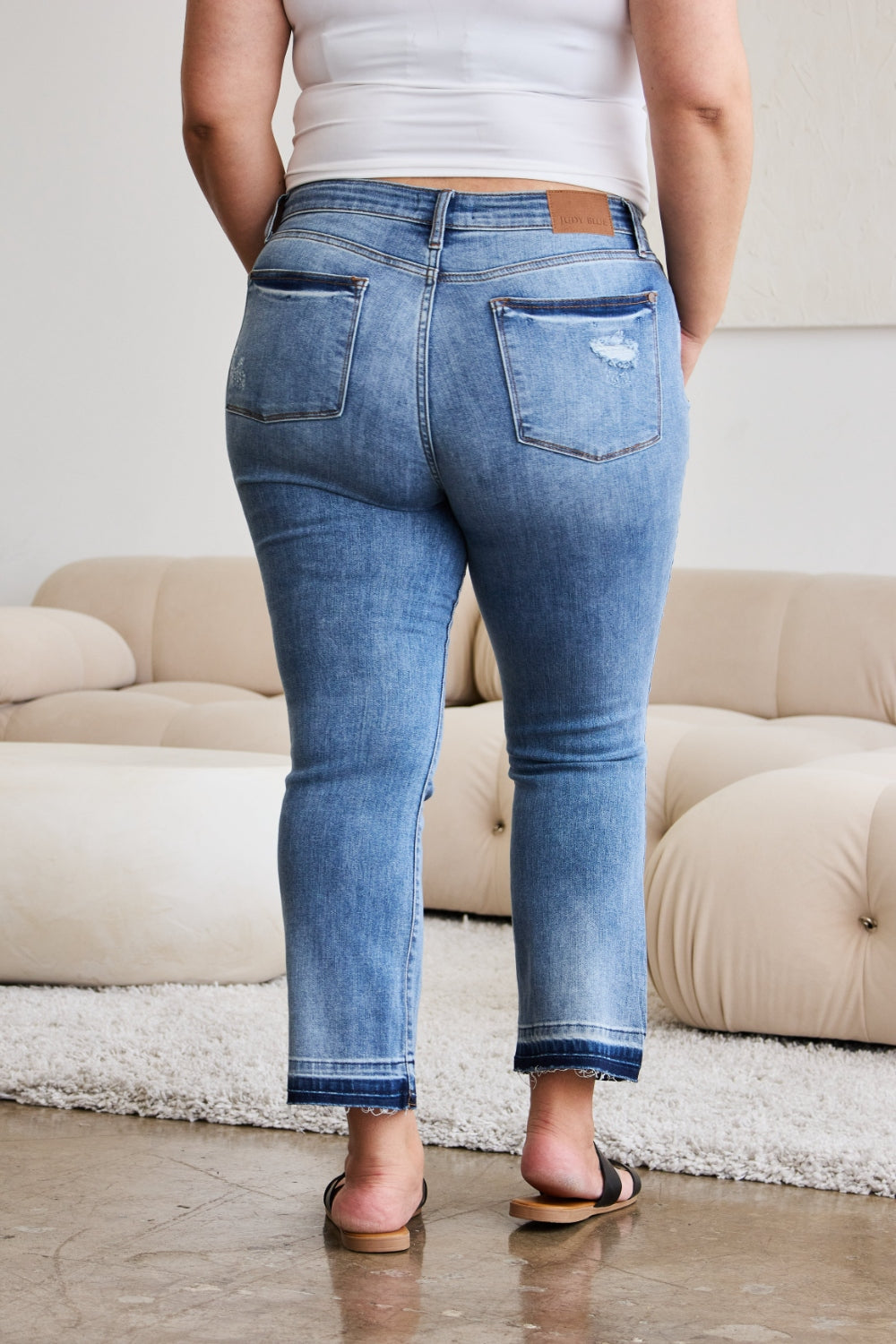 A person modeling Judy Blue Full Size Release Hem Cropped Bootcut Jeans and white heeled sandals stands in a room with light-colored furniture.