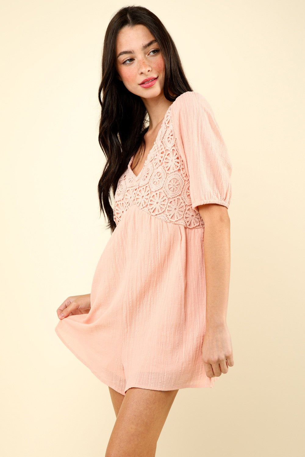 A woman in the VERY J Lace Detail Puff Sleeve Romper with Pockets, featuring light pink fabric and intricate lace detailing around the chest, stands against a plain beige background.