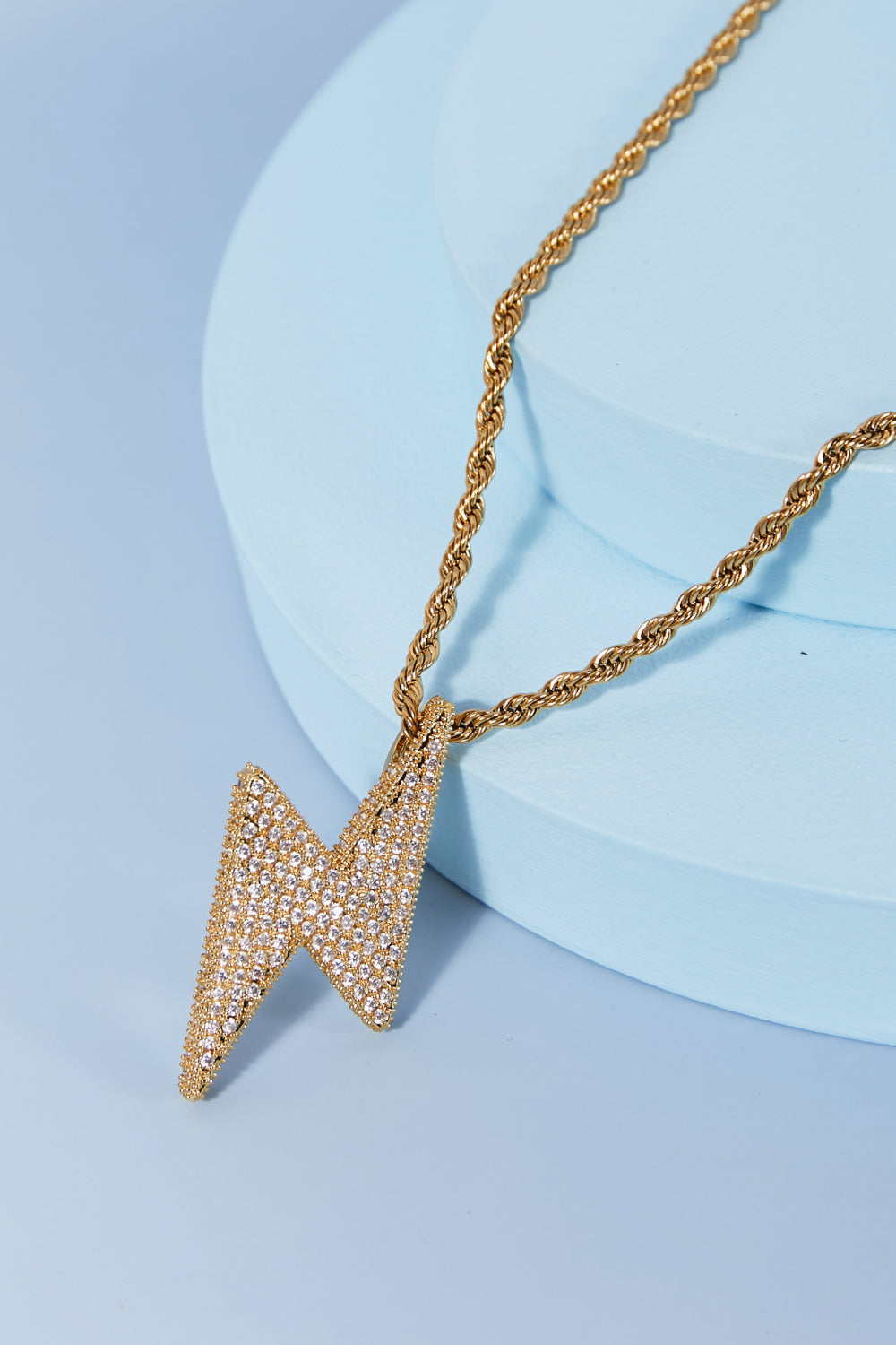 A Brass Lightning Pendant Necklace, featuring a gold twisted chain and a lightning bolt-shaped pendant encrusted with small cubic zirconia stones, displayed on a light blue background.