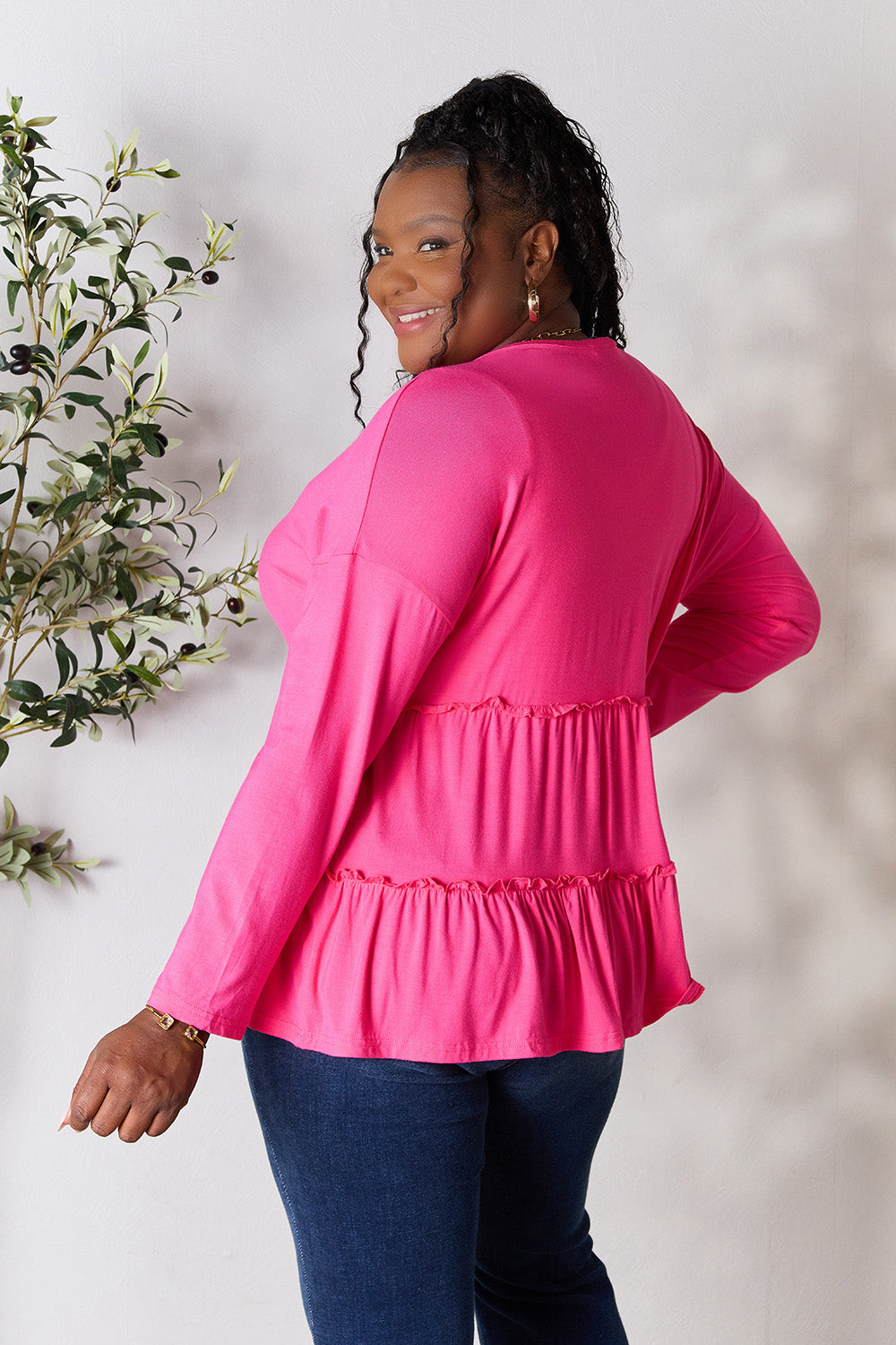 Person stands indoors near a plant, wearing the Double Take Half Button Long Sleeve Ruffle Hem Blouse in gray and blue jeans, smiling while looking to the side. The slightly stretchy fabric of the blouse adds comfort while its buttoned design offers a touch of elegance.