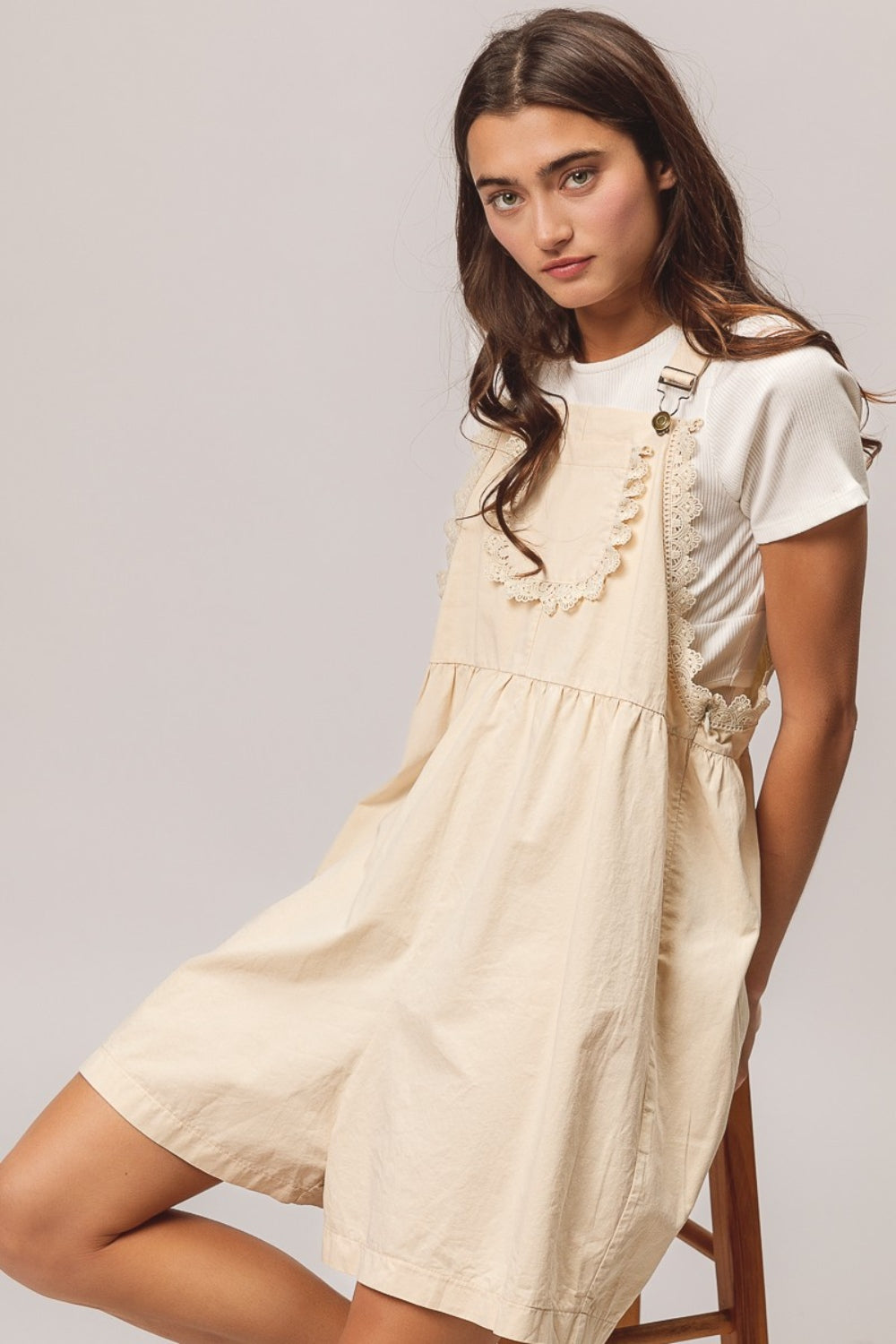 A young woman with long dark hair is sitting on a wooden stool, wearing a white t-shirt paired with the BiBi Back Patch Pocket Lace Trim Rompers—a cream-colored overall dress featuring back patch pockets. With a neutral expression set against a plain light gray background, she effortlessly embodies the perfect addition to any summer wardrobe collection.