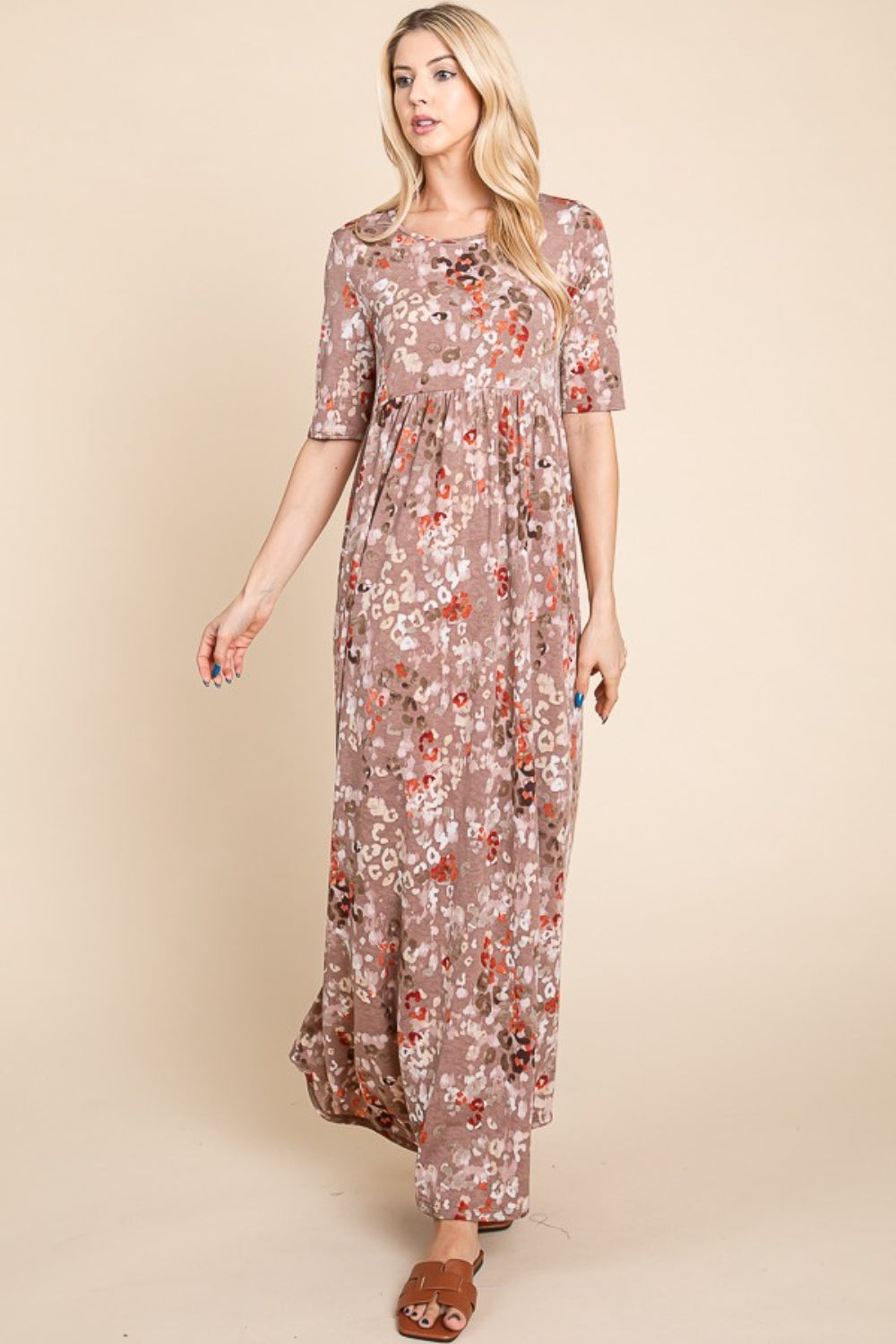 A woman stands against a beige background wearing the light brown, floral BOMBOM Printed Shirred Maxi Dress and brown sandals. She has long blond hair and is looking to her right with one hand slightly raised. The versatile dress offers a flattering fit perfect for any occasion.