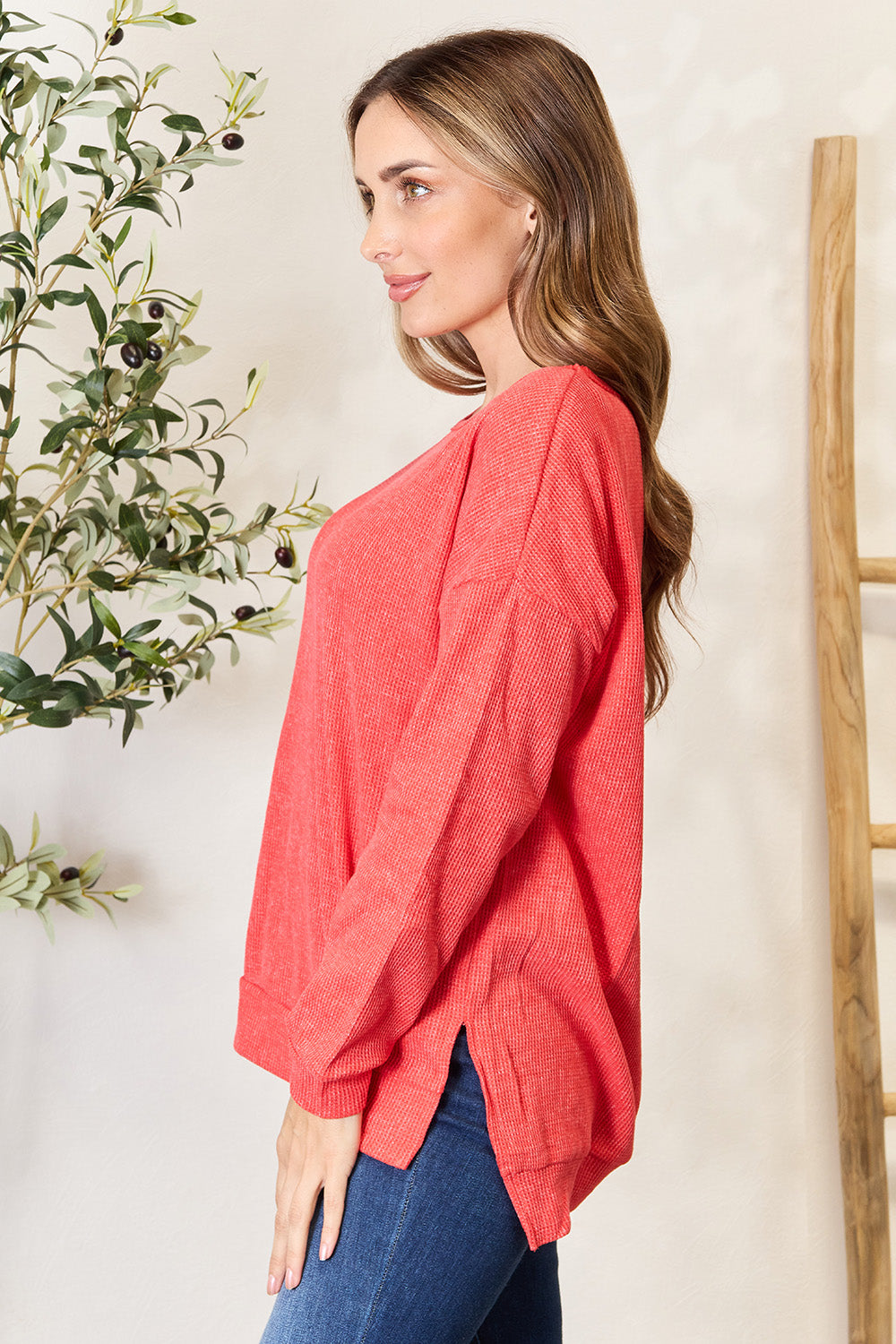 A woman wearing the Basic Bae Round Neck Drop Shoulder Slit Sweatshirt in red and blue jeans stands indoors, smiling and touching her hair, with a decorative plant and wooden ladder in the background.