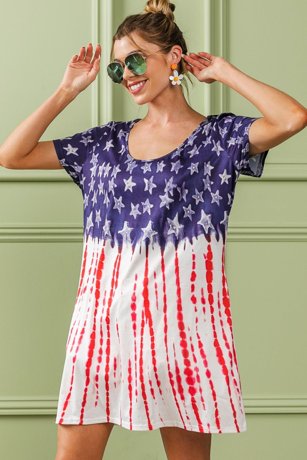 A person wearing the BiBi American Flag Theme Tee Dress, accessorized with sunglasses and earrings, stands against a light green wall with decorative molding.