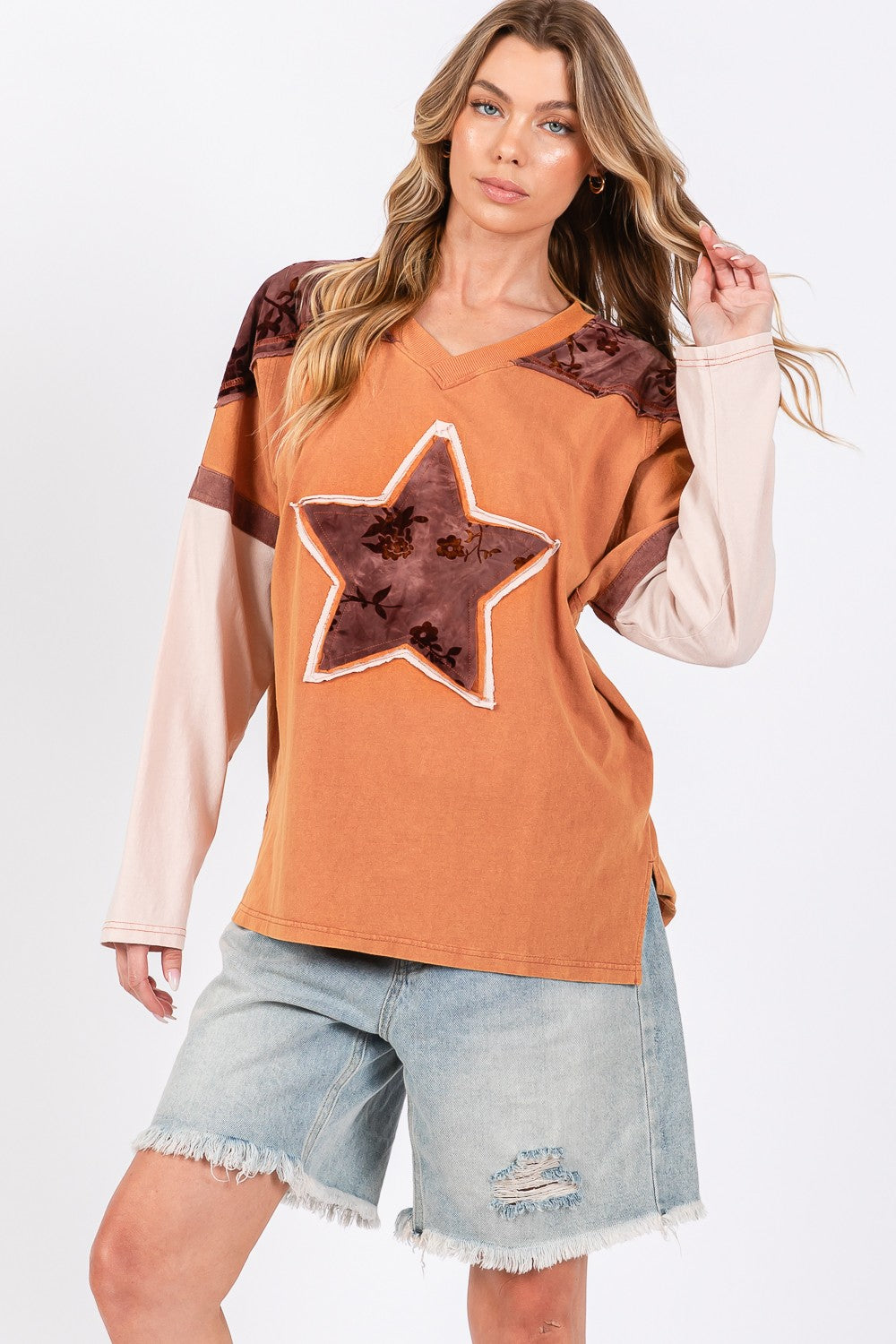A person wearing a beige hat, a SAGE + FIG Star Patch Long Sleeve Color Block T-Shirt in orange, and light blue jeans stands against a plain background.
