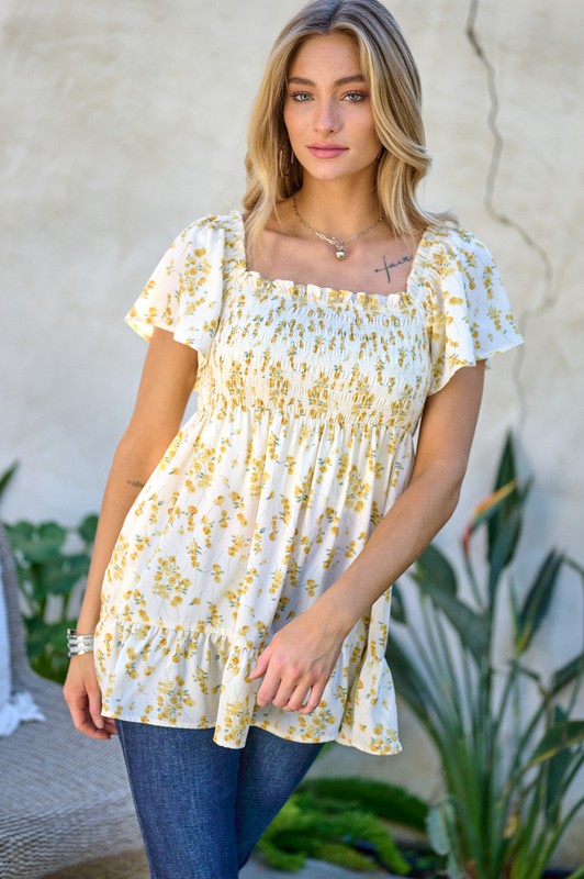 A woman stands outdoors donning a Floral Printed V-Neck Ruffle Top paired with jeans, set against a neutral background, with plants in the foreground, highlighting her fashion-forward wardrobe.